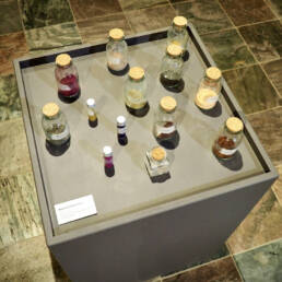 A downward view of a grey plinth which has various small glass bottles of colourful powdered natural dye inside. At Temiskaming Art Gallery, 2023.