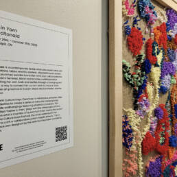 On the left vinyl wall decal that describes the Fables in Yarn project by Alisa McRonald. On the right side, a brightly coloured textile art on canvas at Guelph Civic Museum, 2023.