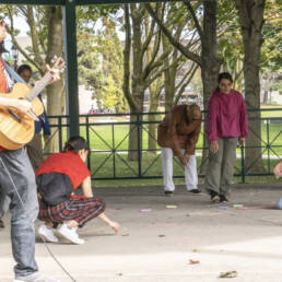 People gather under a gazebo at The Esplanade Park, during Mushtari Afroz’s Choreo-xperience Public Spaces, 2023. On the left a man in a red shirt plays a guitar, in the centre a few squat and draw on the cement with chalk and stretch.