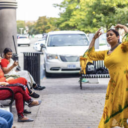Several people sit on green metal benches in a row, watching a performer in a yellow dress dance on the right at Mushtari Afroz’s Choreo-xperience Public Spaces, 2023.