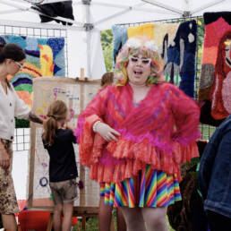 A drag performer with pastel rainbow coloured hair, a rainbow dress, and bright pink/red sheer coat, and white heart shaped sunglasses stands in front of Alisa McRonalds tent at the outdoor Queen West Art Crawl 2023.