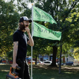 Owen Marshall’s stands in front of his flag installation; two green flags, top: “MY LIFE! HAS PURPOSE!” and bottom: “AGAIN!”, 2023.