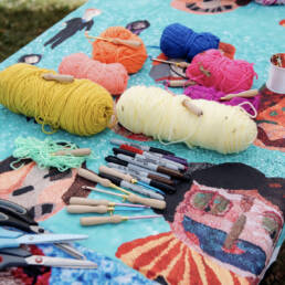 A close-up image of a table covered a teal fabric with colourful yarn, needle punches, and various craft tools on top, 2023.