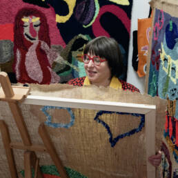 A close up of a woman working on a large stretched canvas with yarn on an easel, surrounded by large colourful wall hangings