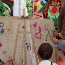 Two adults and two children work on a needle punch canvas, with colourful yarn at Alisa McRonald’s tent at the Queen West Art Crawl 2023.