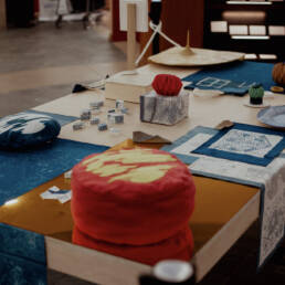 An overview image of the project’s artefacts from Myung-Sun Kim’s exhibition Rituals for Belonging at Toronto Public Library – Lillian H. Smith Branch, 2023. On a light coloured wooden table sits a variety of items, including; swatches of blue fabric, small blue and white boxes, small cushions, and folded paper.