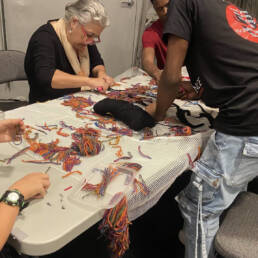 Several people work around a table on looping and weaving a textile piece together at a Kevin A. Ormsby workshop, 2023.