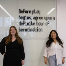 Chloé Ouellet-Riendeau (left) and Juliane Foronda (right) stand in front of a glass wall with black vinyl lettering that reads as “Before play begins, agree upon a definite hour of termination.” from Foronda’s project NOTES ON PLAy at the Ottawa Art Gallery, 2023.