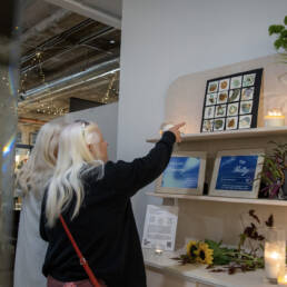 Two people look at and point towards a shelf that was information about and work by Betty Carpick, along with flowers and candles at Co. Lab Gallery and Arts Centre, 2023.