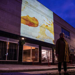 Betty Carpicks stands in front of a projection of her artwork at the Downtown Fitness building in Thunder Bay, 2023.