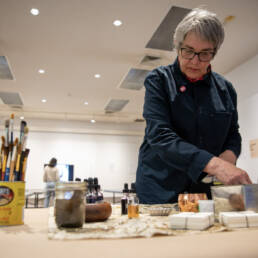 Betty Carpick stands at a table looking down at the brushes and inks as she works at her workshop, Thunder Bay Art Gallery 2023.