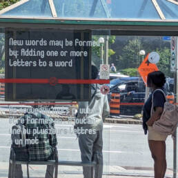 Three people wait inside a glass bus stop in Ottawa that is decorated with white vinyl words from Juliane Foronda’s project NOTES ON PLAY. The vinyl reads “New words may be formed by: Adding one or more letters to a word” in english and below in french, 2023