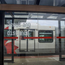A glass bus stop in Ottawa with white vinyl words from Juliane Foronda’s project NOTES ON PLAY that reads as “Scout any distance,” 2023.