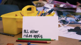 A close-up of a table covered in crafting supplies, a white card that reads “All other rules apply.” leans against a yellow pencil caddy at Juliane Foranda’s workshop, 2023.