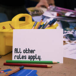 A close-up of a table covered in crafting supplies, a white card that reads “All other rules apply.” leans against a yellow pencil caddy at Juliane Foranda’s workshop, 2023.